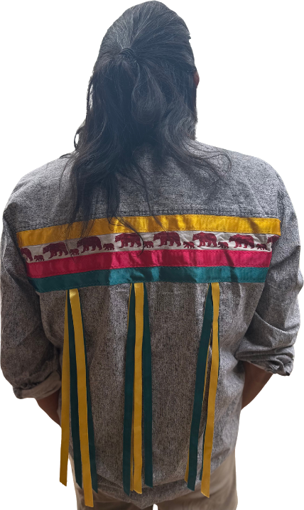 Standing Bears from behind, wearing his ribbon shirt: bands of gold, white with bears walking across it, crimson and teal, with ribbons hanging down the back.
